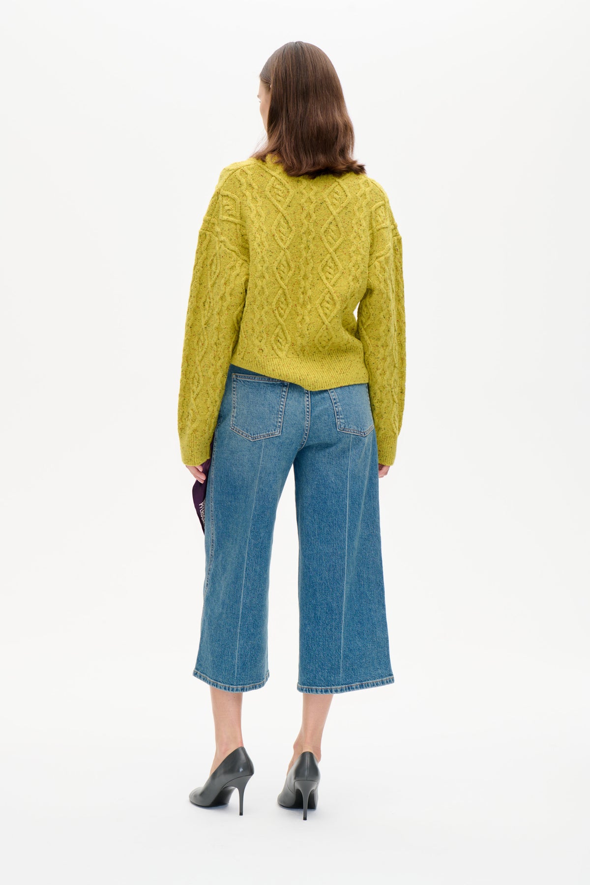 Back view of a model wearing a yellow-green flecked cable-knit sweater with blue cropped wide-leg jeans.