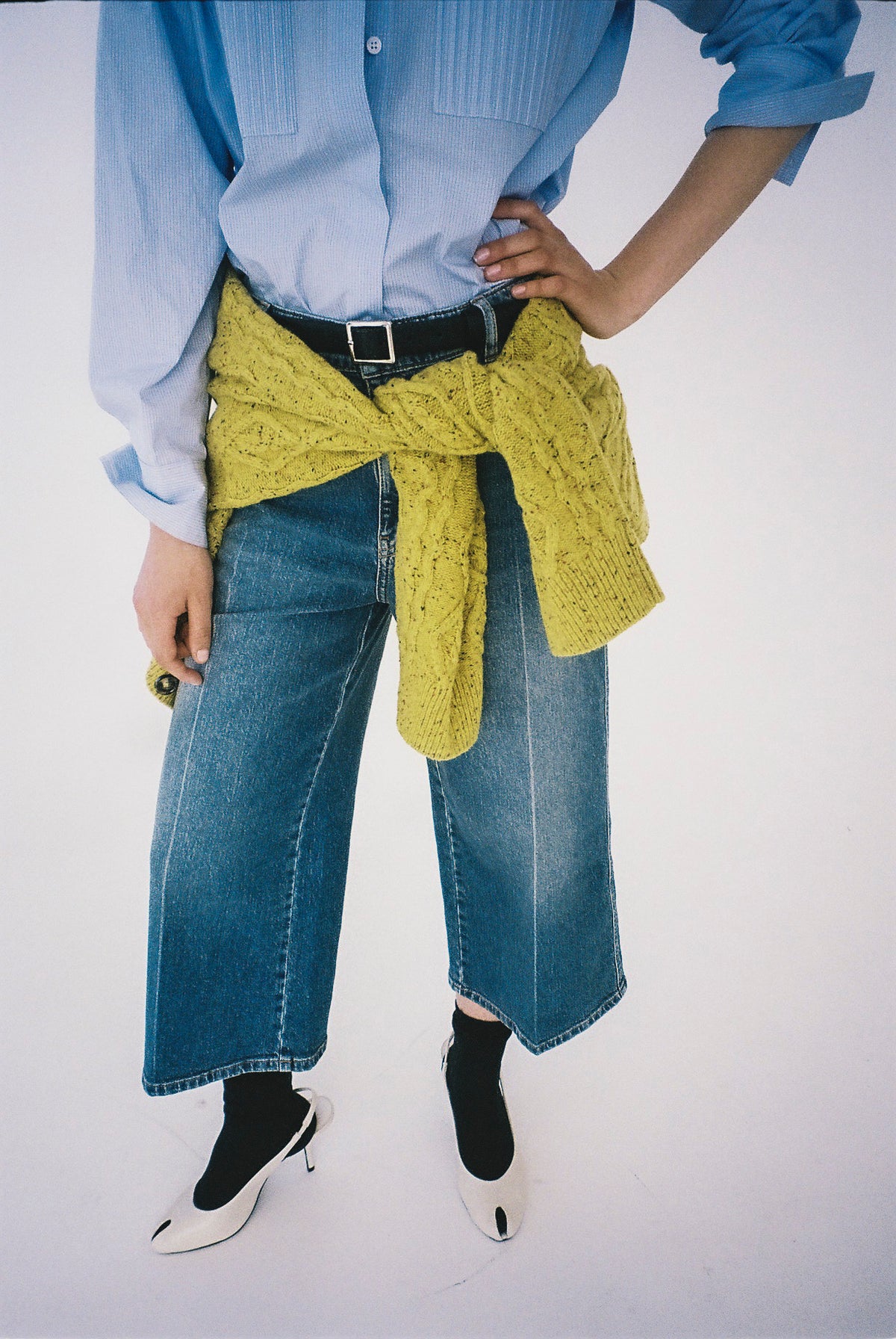 Cropped campaign view of a light blue button-up shirt, wide-leg blue jeans, and a mustard yellow sweater tied at the waist.