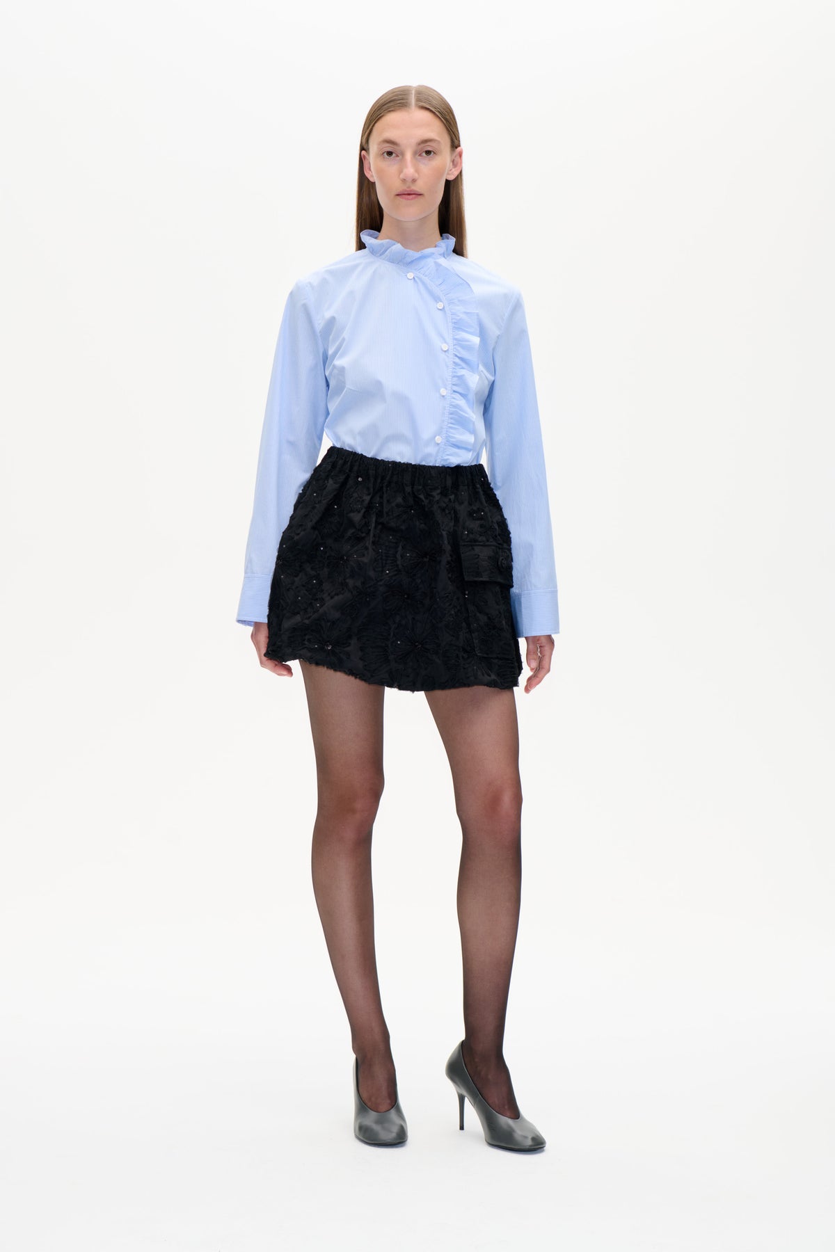 Front-facing model in a pale blue pinstripe shirt with a ruffled collar and asymmetric ruffle placket, paired with a black textured mini skirt.
