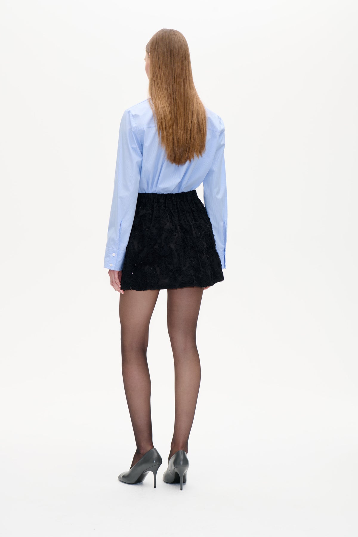 Back view of a model wearing a pale blue pinstriped shirt with a black textured floral mini skirt.