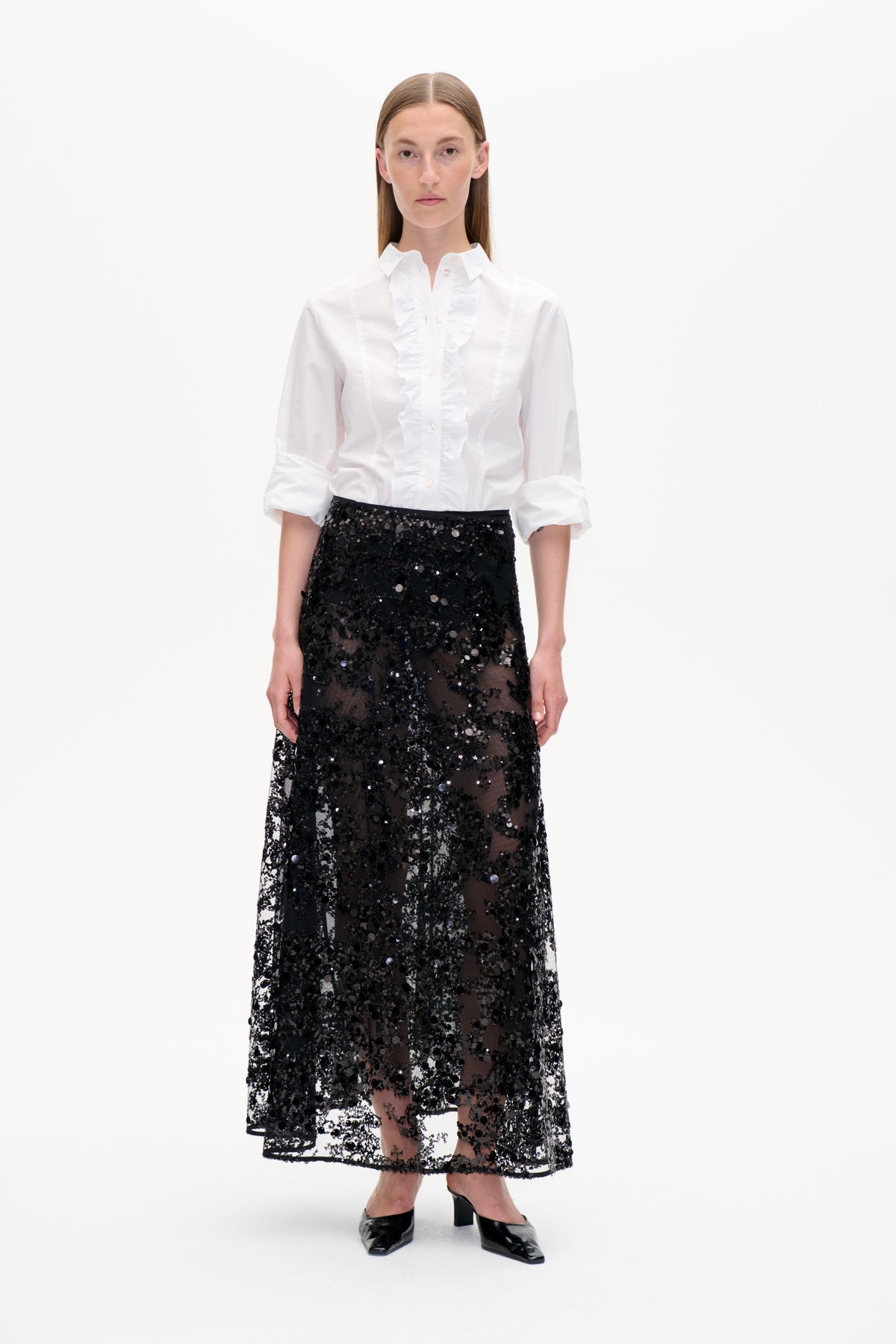 Front view of a model wearing a white ruffled button-up shirt and a sheer black sequin midi skirt against a white background.