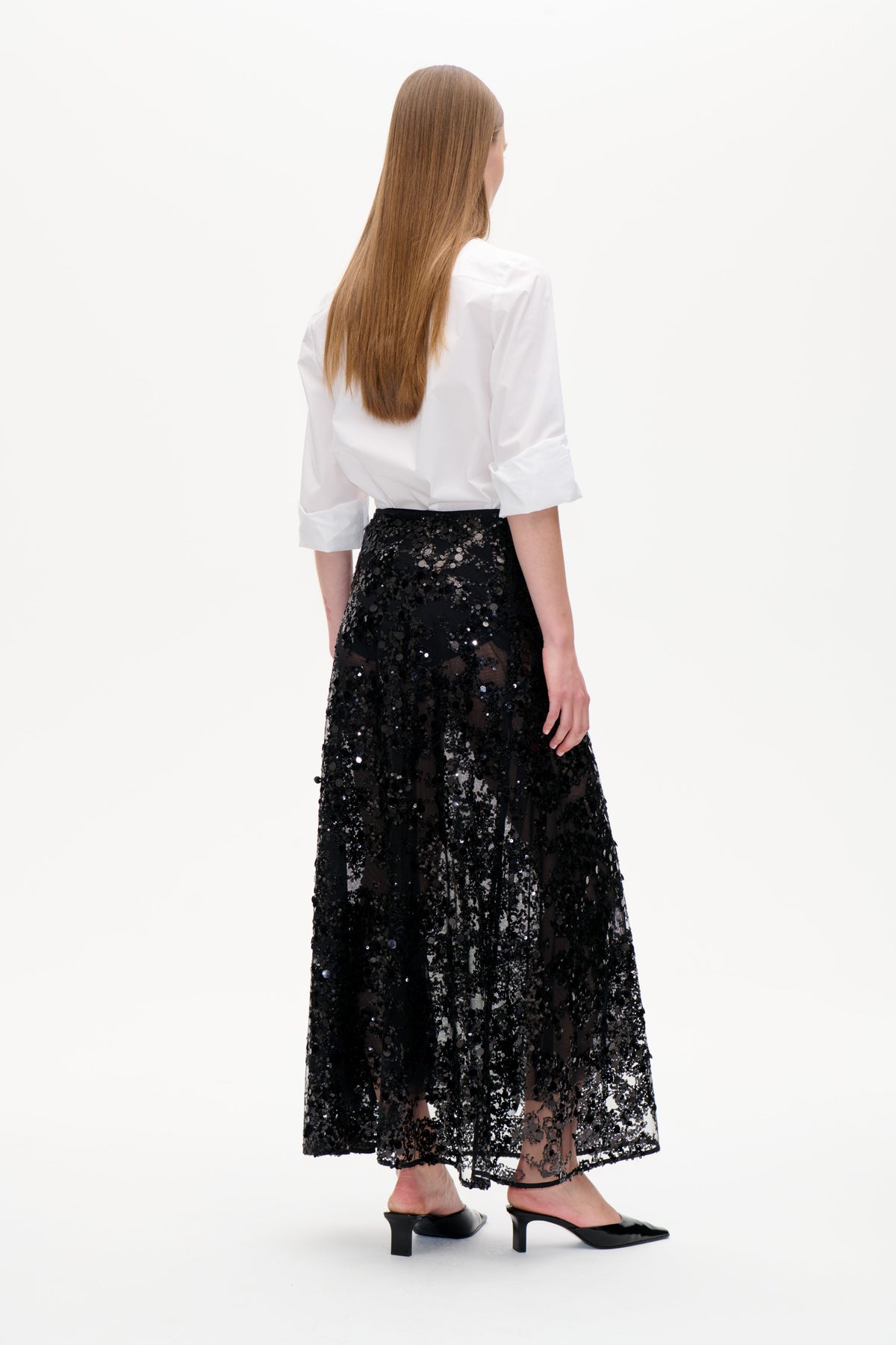 Back view of a model wearing a white shirt and a black sheer midi skirt densely covered in sequins.
