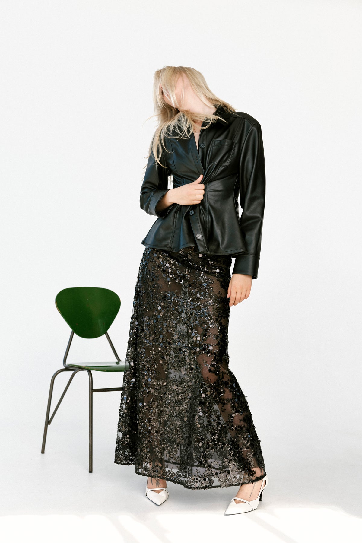 Campaign portrait of a model in a black button-front jacket and sheer black sequin midi skirt, beside a green chair against white.