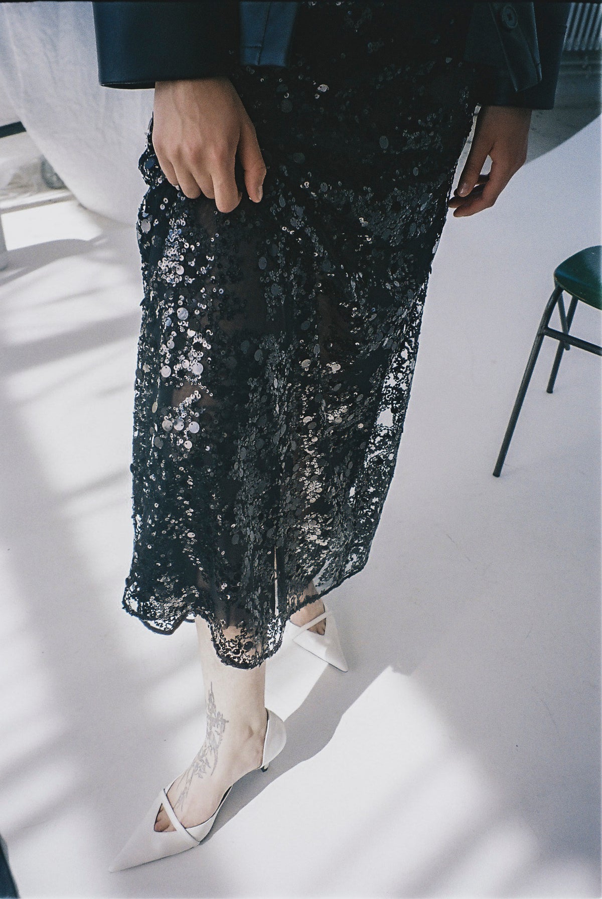 Close-up campaign view of a model wearing a black sheer skirt covered in silver sequins and white pointed heels.