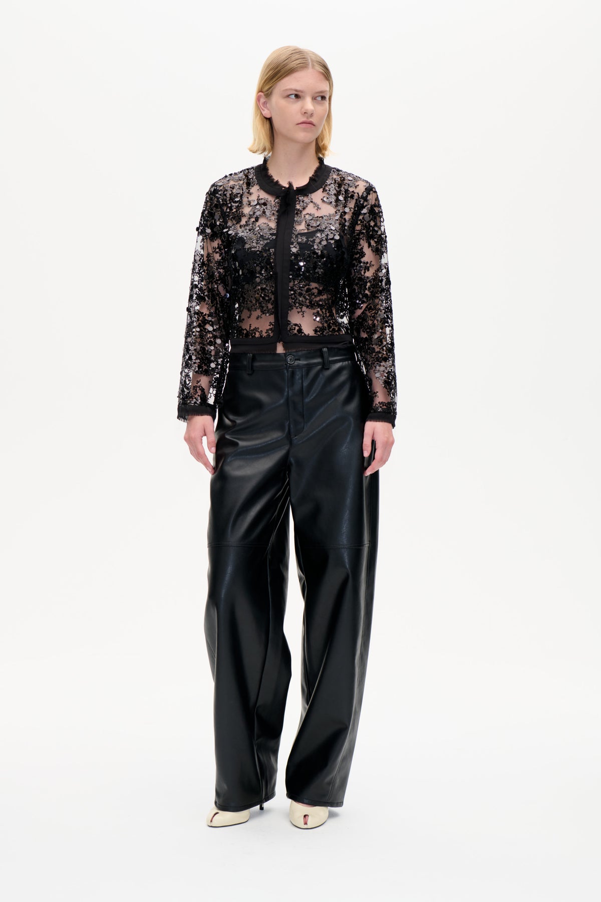 Model wearing a sheer black sequin blouse with a tied trim collar and wide-leg black trousers, shown front-on against a white studio background.