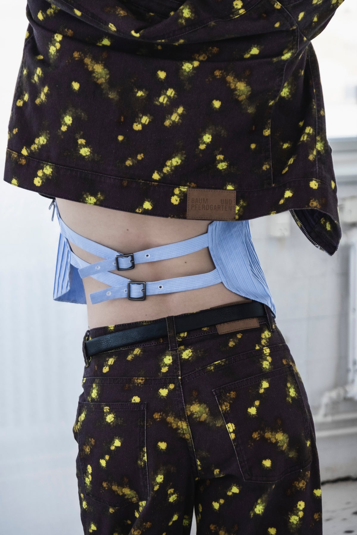 Close-up campaign styling of a dark denim jacket and matching trousers with yellow-green floral print over a blue striped cutout shirt with buckle straps.