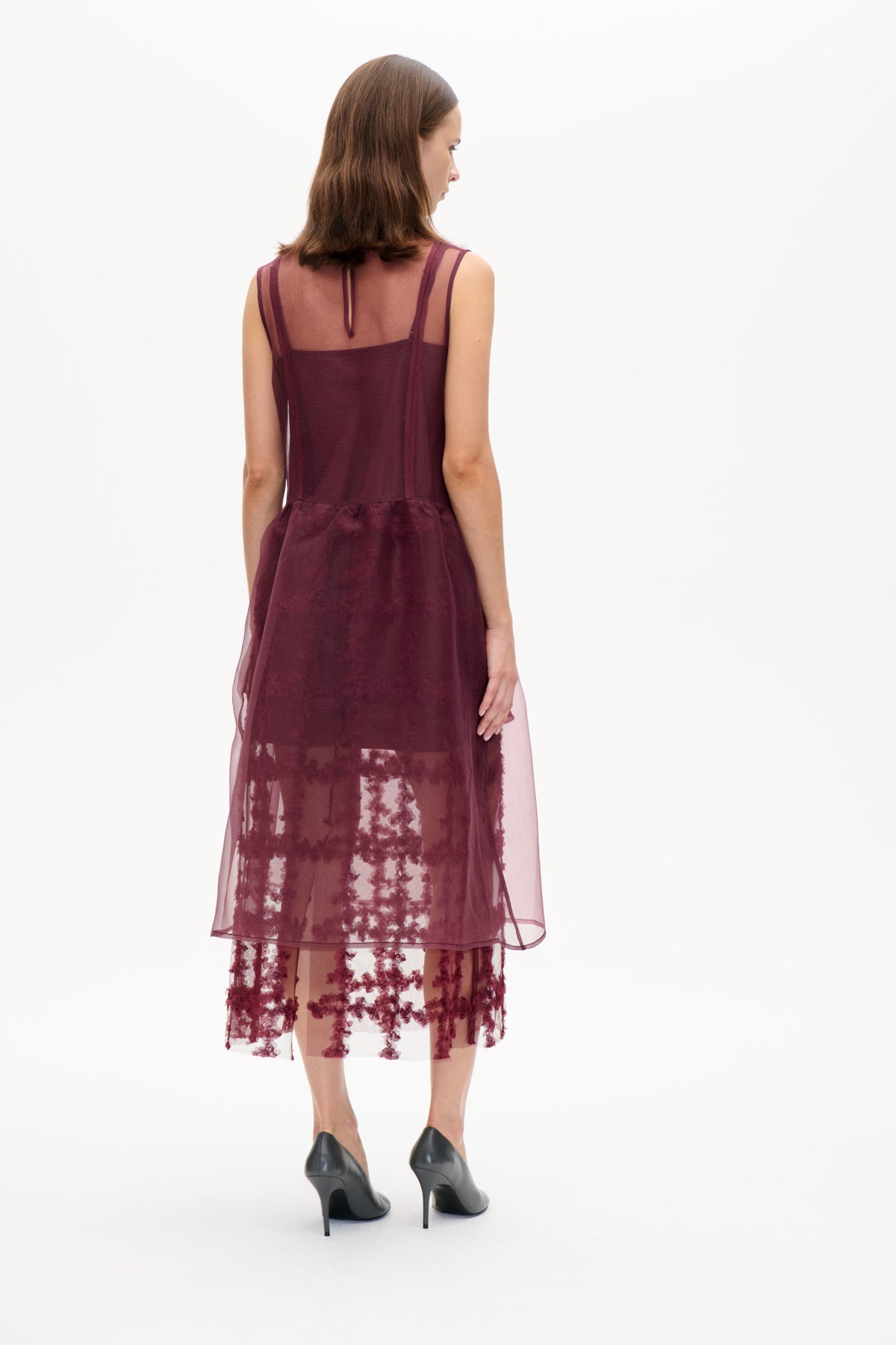 Rear view of a model in a burgundy sleeveless layered dress with a sheer back panel and floral-patterned transparent skirt
