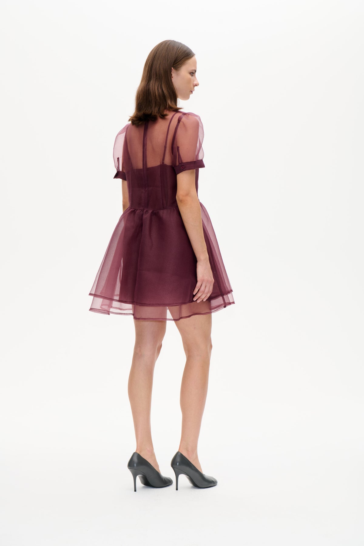 Model in a plum mini dress with sheer mauve puff sleeves and a layered transparent skirt, shown from a rear three-quarter angle.