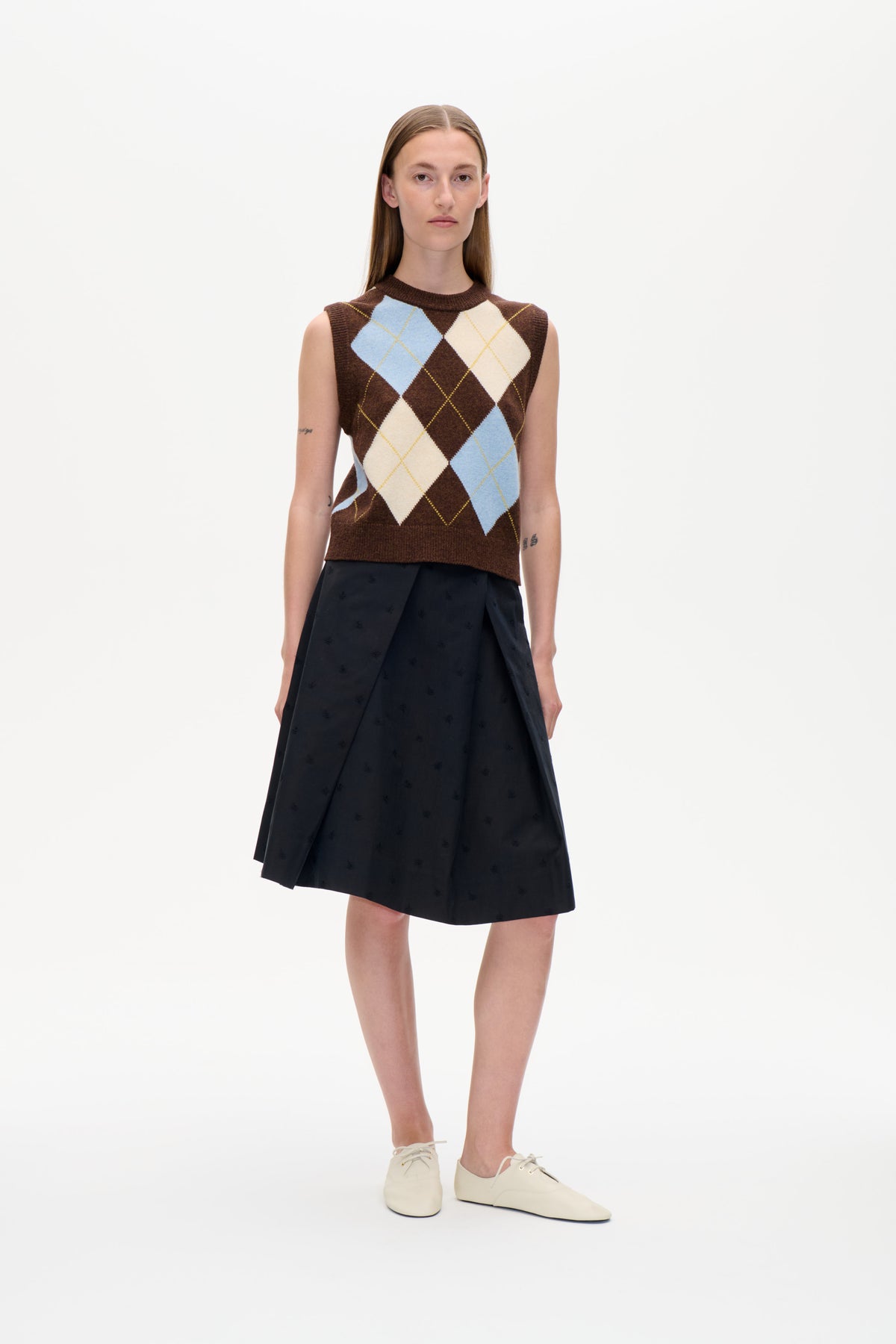 A model stands against a white background wearing a brown sleeveless knit vest with blue and cream argyle diamonds and a black patterned skirt.