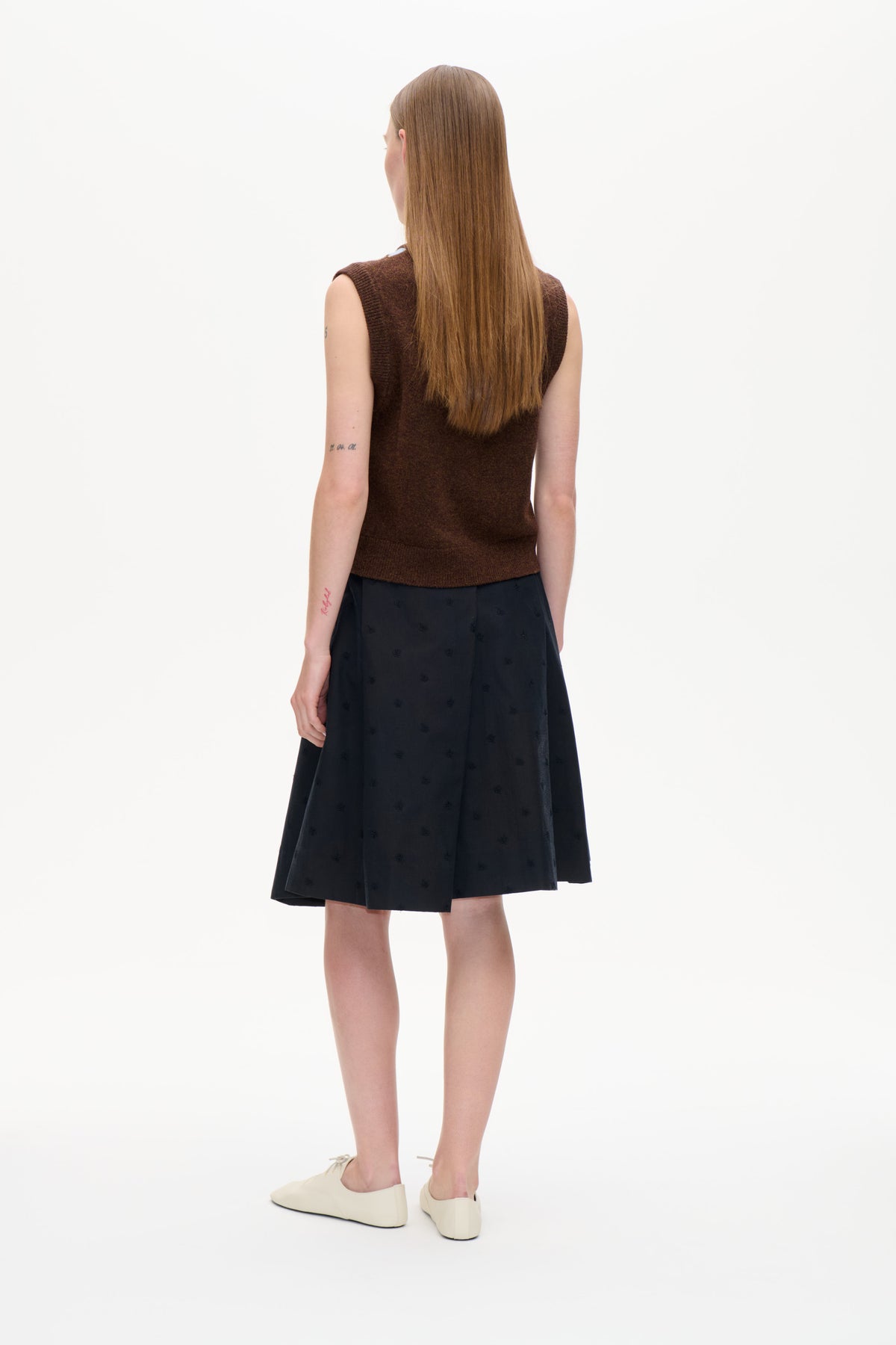 Back view of a model wearing a dark brown sleeveless knit top, black patterned midi skirt and cream flats.