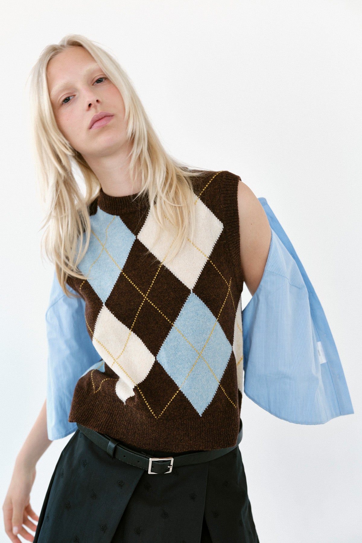 Model wearing a brown sleeveless knit vest with blue and cream argyle diamonds over a light blue shirt.