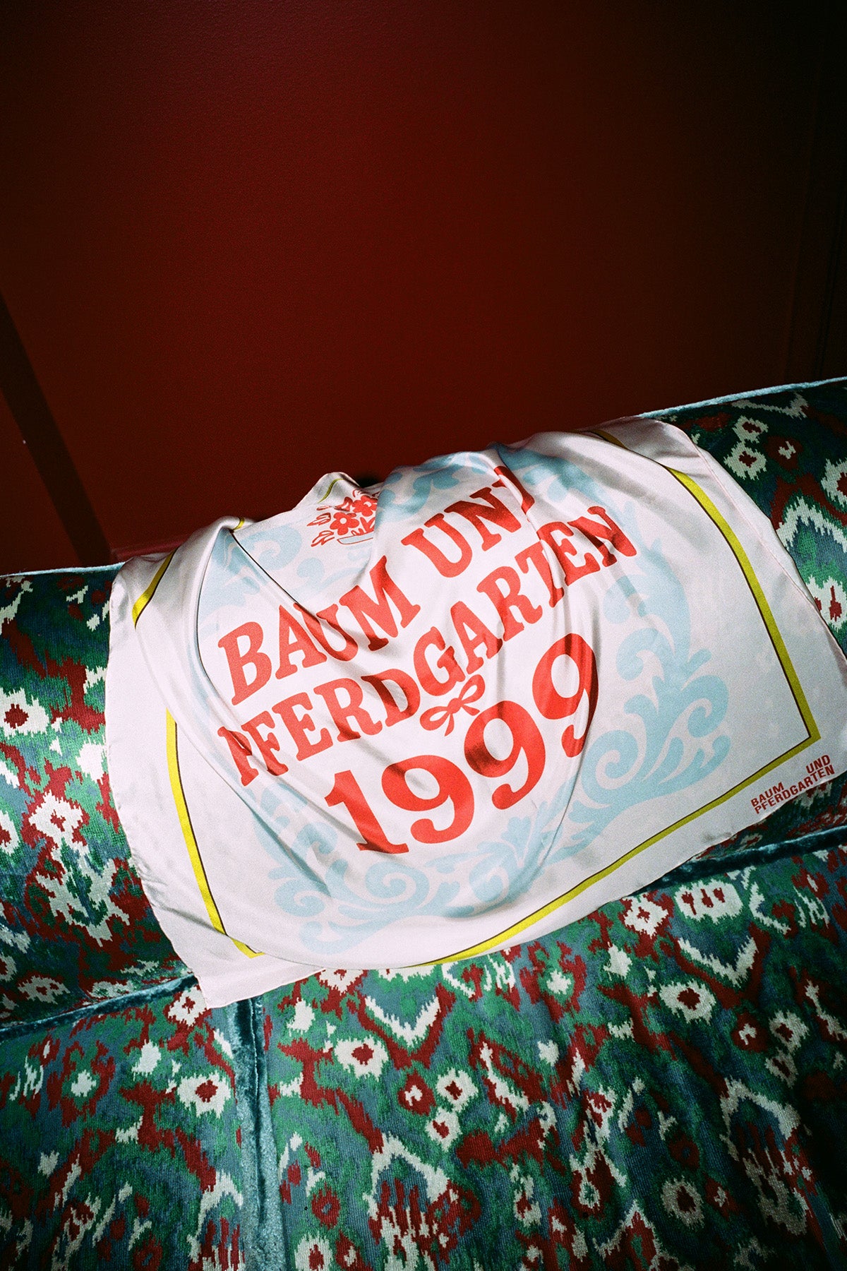 Printed square scarf draped over a patterned chair, with pale blue scrollwork, yellow edging and red “Baum und Pferdgarten 1999” lettering.