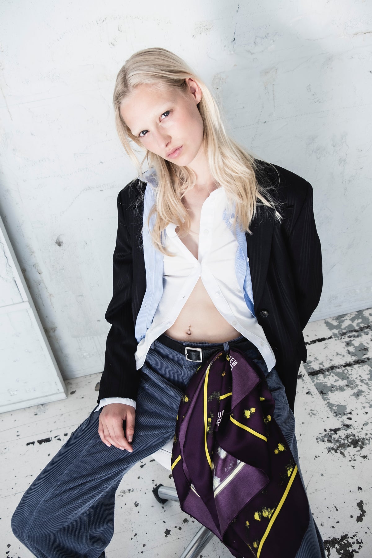 Campaign portrait of a model in a black blazer, white collared shirt and pale blue pinstriped vest, with a purple floral scarf across dark blue trousers.