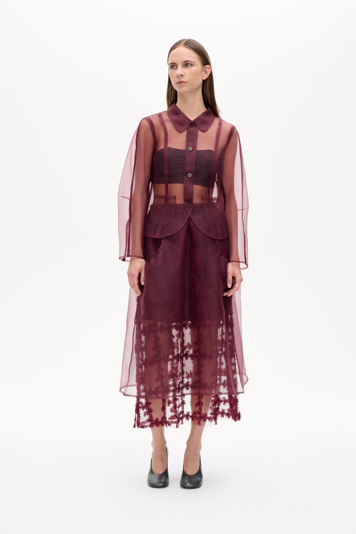 Model draagt een doorschijnende pruimkleurige tuniek met knoopsluiting aan de voorkant, verticale strepen, ronde kraag en opgestikte zakken, over een donkere top en een rok met bloemenprint