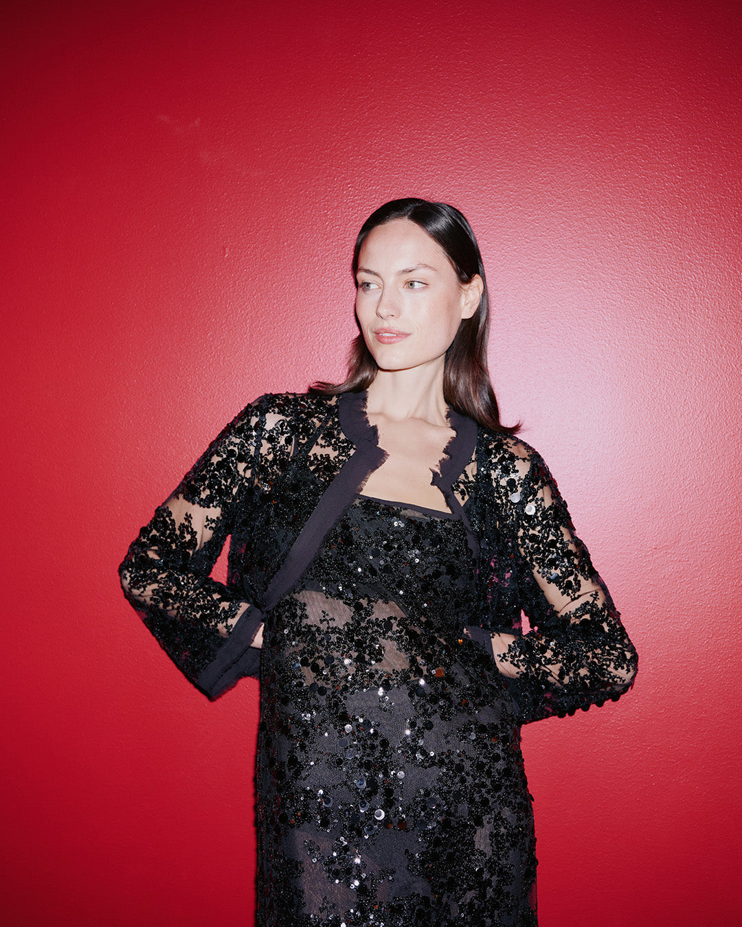 Model wearing a black sequin dress with a sheer floral lace, long-sleeved jacket against a red wall